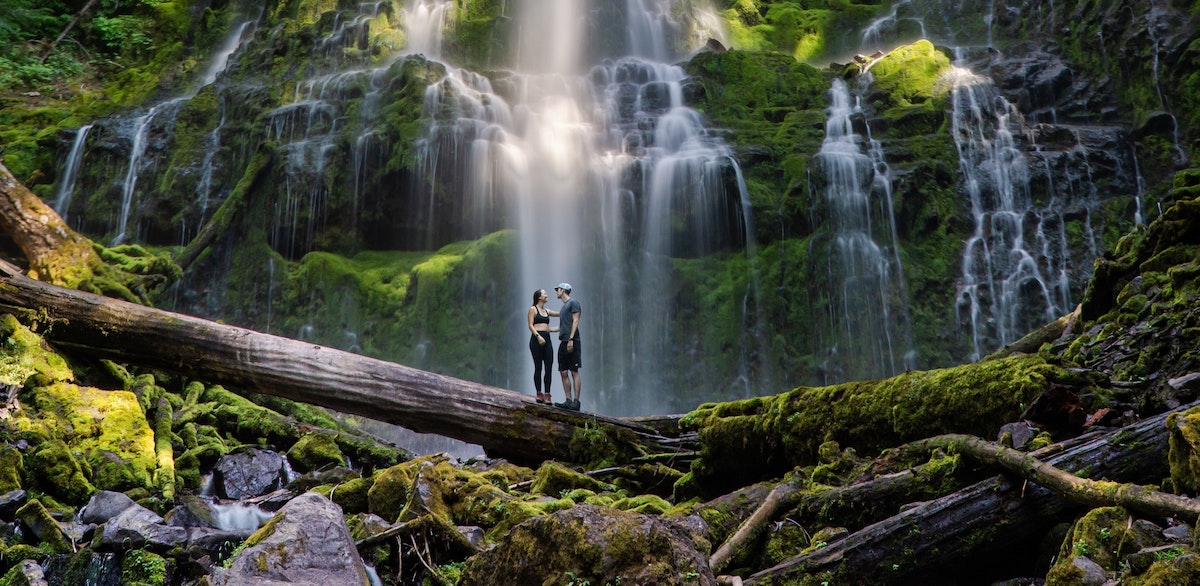 Couple standing on a fallen log framed against a misty waterfall covered in moss in an Oregon forest